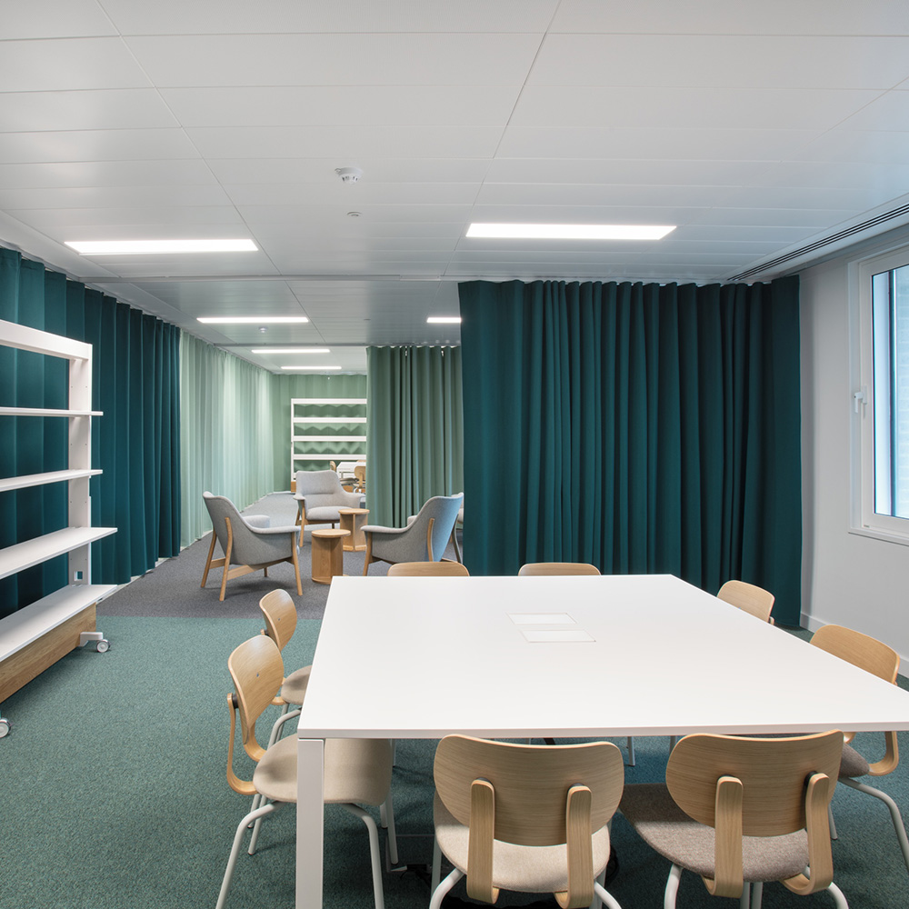 Bureau & espaces de travail 18 Des rideaux acoustiques bleu pétrole et vert servant de séparateurs d'espace dans une salle de formation ou une bibliothèque lumineuse, équipée de chaises en bois et d'étagères blanches