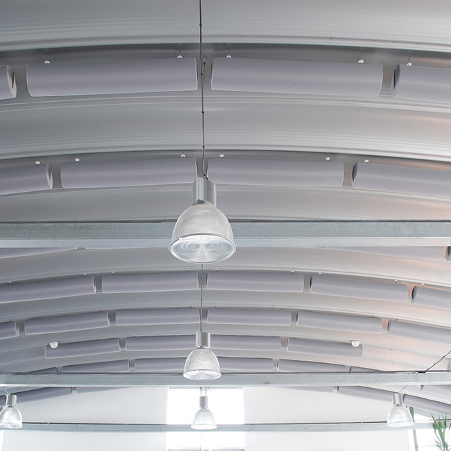 Absorbeur cylindrique acouFoam NoFlame* 7 Des absorbeurs cylindriques gris, montés transversalement entre les arceaux métalliques d'un plafond industriel voûté équipé de luminaires suspendus