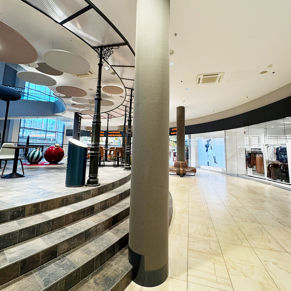 Industria & commercio 16 In primo piano, colonne rivestite con un materiale fonoassorbente grigio-marrone e, sullo sfondo di un centro commerciale, pannelli fonoassorbenti rotondi di colore rosa, grigio e bianco