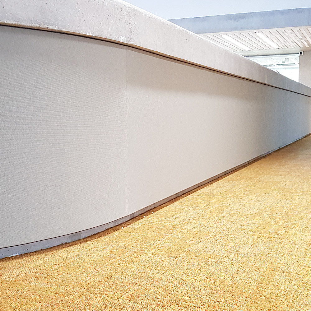 Acustica & rumore 15 Rivestimento murale in tessuto grigio chiaro senza giunti, posato su una parete curva in un grande atrio con moquette gialla e soffitto in cemento a vista