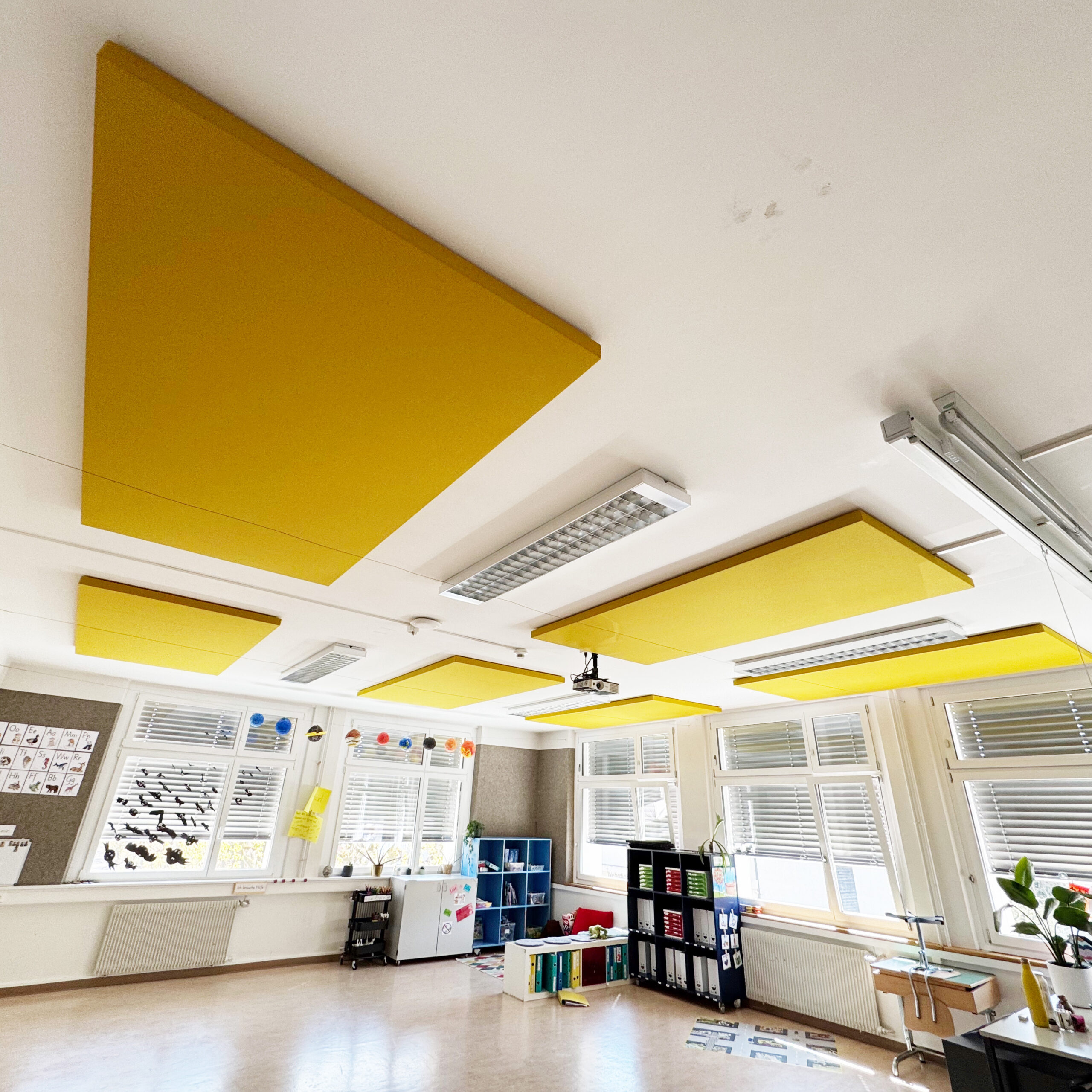 Scuola & istruzione 14 Grandi pannelli acustici gialli fissati direttamente al soffitto bianco di un’aula, con un proiettore e arredi scolastici colorati